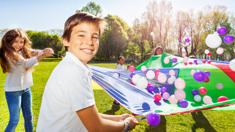 Junge hält eine Ecke eines bunten Tuches fest, auf dem Bälle herumfliegen. Er ist der Kamera zugewandt und lächelt.