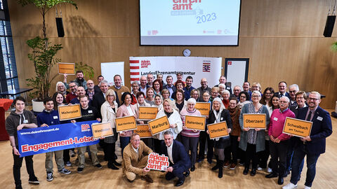Staatsminister Axel Wintermeyer gemeinsam mit allen neuen Engagements-Lotsinnnen und Lotsen. Die Lotsinnen und Lotsen halten Bilder von Ortsschildern. 