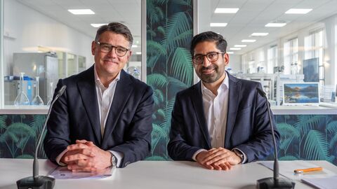 Zwei Herren im Anzug lachen in Kamera. Vor beiden steht ein Mikrofon.