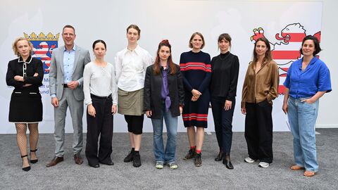 Auf dem Bild sind zu sehen v.l.nr.: Catrine Val (Hauptstipendiatin), Christoph Degen (Staatssekretär im Hessischen Ministerium für Wissenschaft und Forschung, Kunst und Kultur), Laura Stellacci (Nachwuchsstipendiatin), Eva Tatjana Stürmer (Arbeitsstipendiatin), Sopo Kashakashvili (Arbeitsstipendiatin), Julia Mihály und Sina Ahlers (teilen sich ein Hauptstipendium), Sophia Solaris (Arbeitsstipendiatin) und Eleonora Herder (Arbeitsstipendiatin). Staatssekretär Degen mit acht Frauen stehen in einer Reihe. Auf der Wand hinter ihnen ist das Hessen Wappen aufgedruckt.