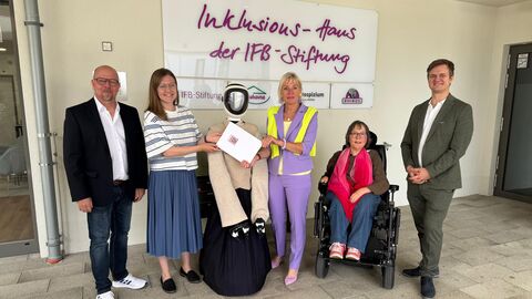Pilotprojekt Einsatz von Robotik  Mehrere Menschen stehen mit einem Roboter vor einer Wand und lachen in die Kamera. Eine Person sitzt im Rollstuhl